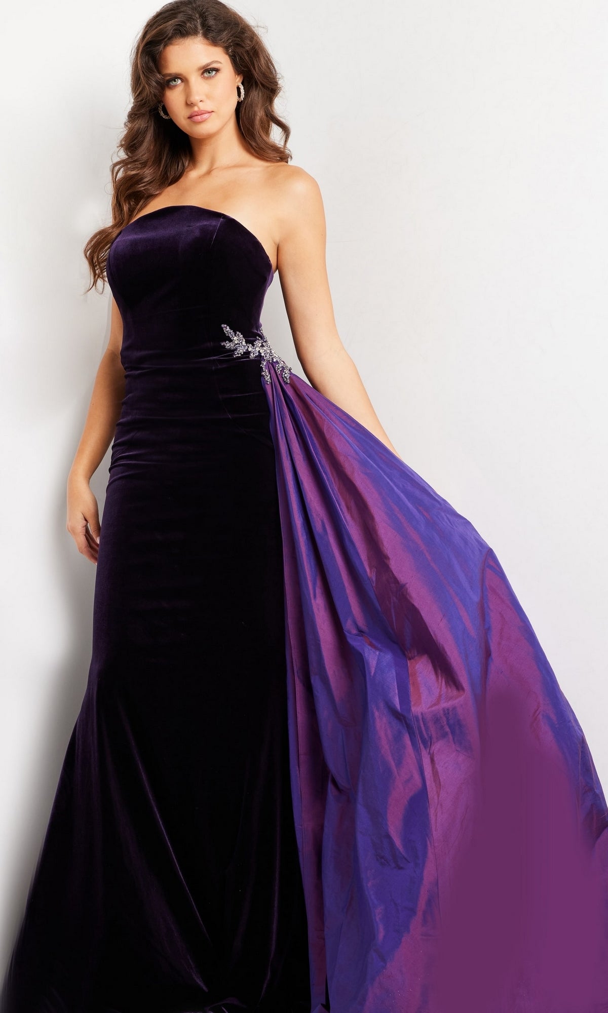 Woman wearing the Jovani Strapless Long Velvet Prom Dress 26116, a floor-length dark purple gown featuring an attached shiny purple drape and silver embellishment.